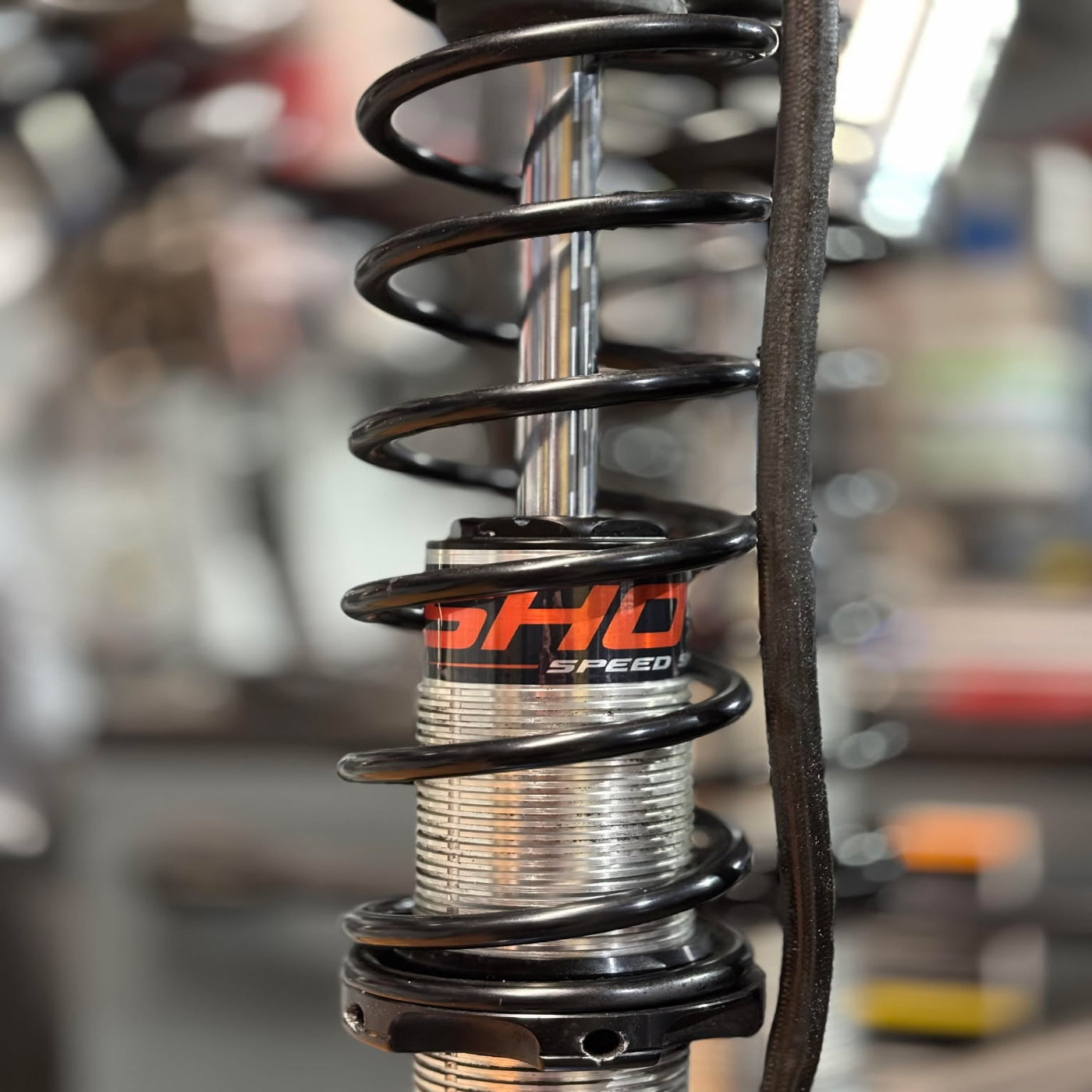 Close-up of a shock absorber with 'Sho-Me Speed' branding in a blurred workshop setting